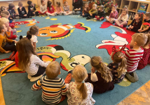 Dzieci siedzą na dywanie słuchając nauczyciela.